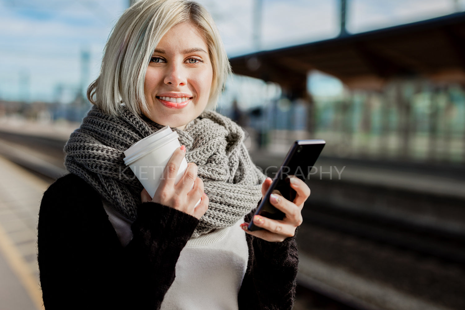 Woman Holding Coffee Cup And Mobile Phone At Train Station