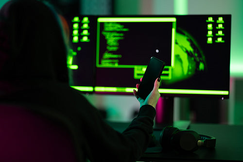 Hacker holding smartphone in front of dual screens with coding interface