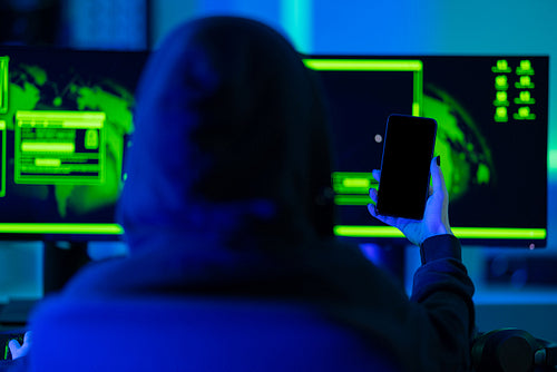 Hacker using smartphone in front of dual monitors with green glow