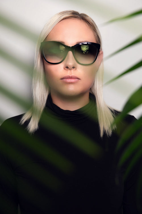 Close-up of woman with sunglasses hiding behind tropical palm leaves