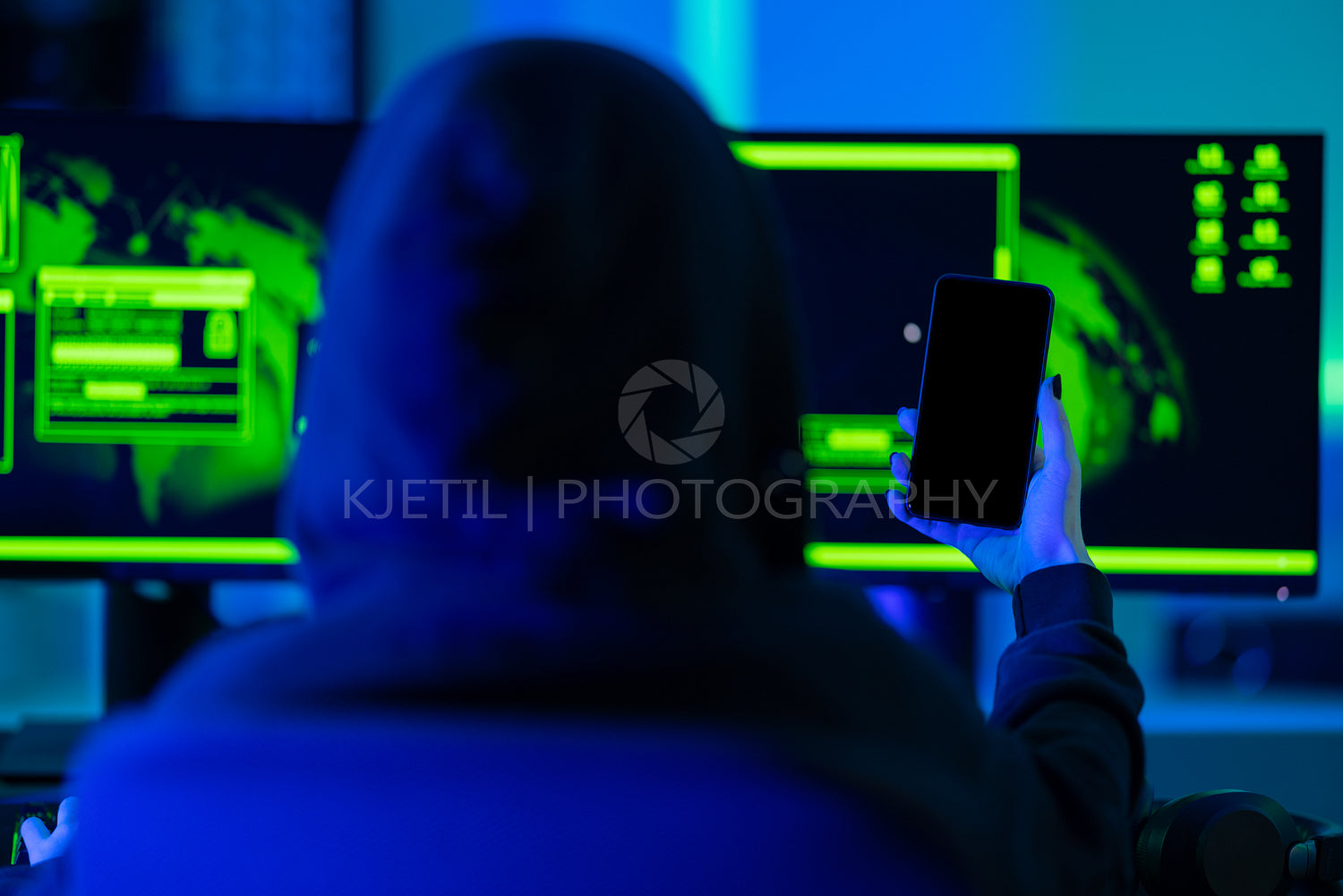 Hacker using smartphone in front of dual monitors with green glow