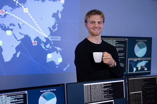 Confident male Cybersecurity Analyst with coffee cup in large Cyber Security Operation Center SOC handling Threats