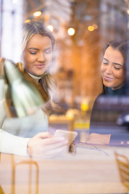 Two Female Friends Looking At Smartphone In Cafe seen through window