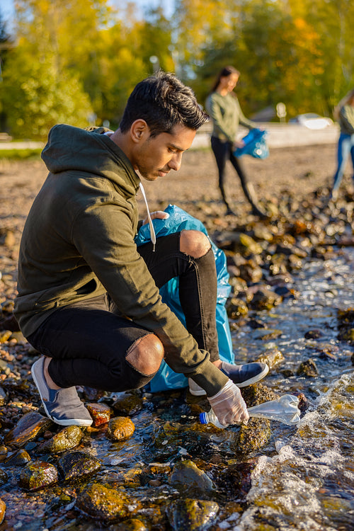 Young volunteer man crouching while picking up garbage on shore