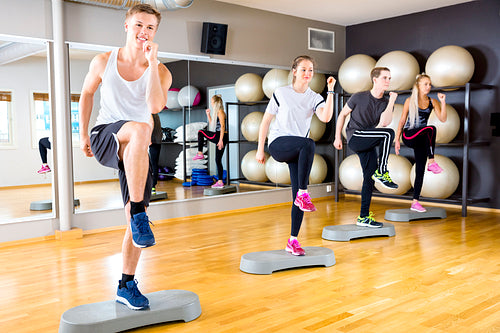 Smiling group raising legs on step platforms at fitness gym