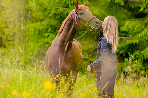 Blonde woman standing in a meadow kissing her arabian horse