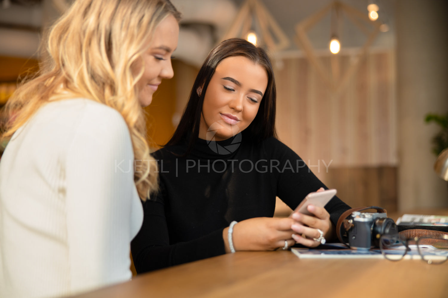 Two Smiling and Beautiful Young Friends Looking At Smartphone In Cafe