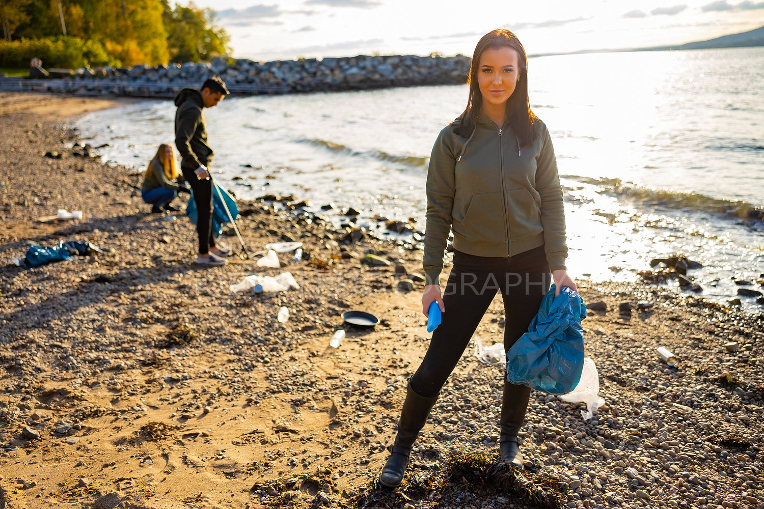 Smiling young woman cleaning beach with volunteers during sunset