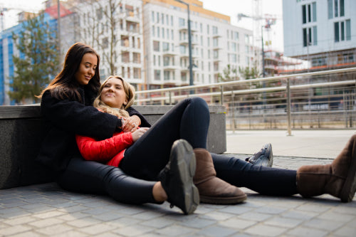 Happy Best Friends Embracing And Sitting On The Ground In City