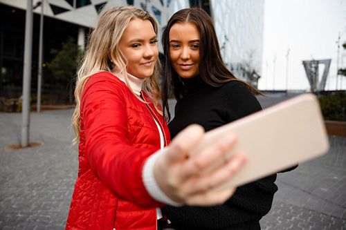 Two Smiling Beautiful Friends Taking Selfie Through Smartphone In City