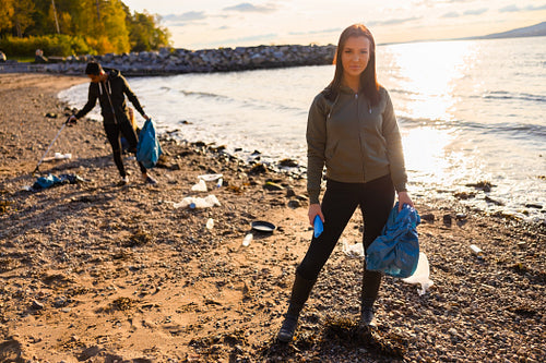 Serious young woman cleaning beach for plastic with volunteers during beautiful sunset