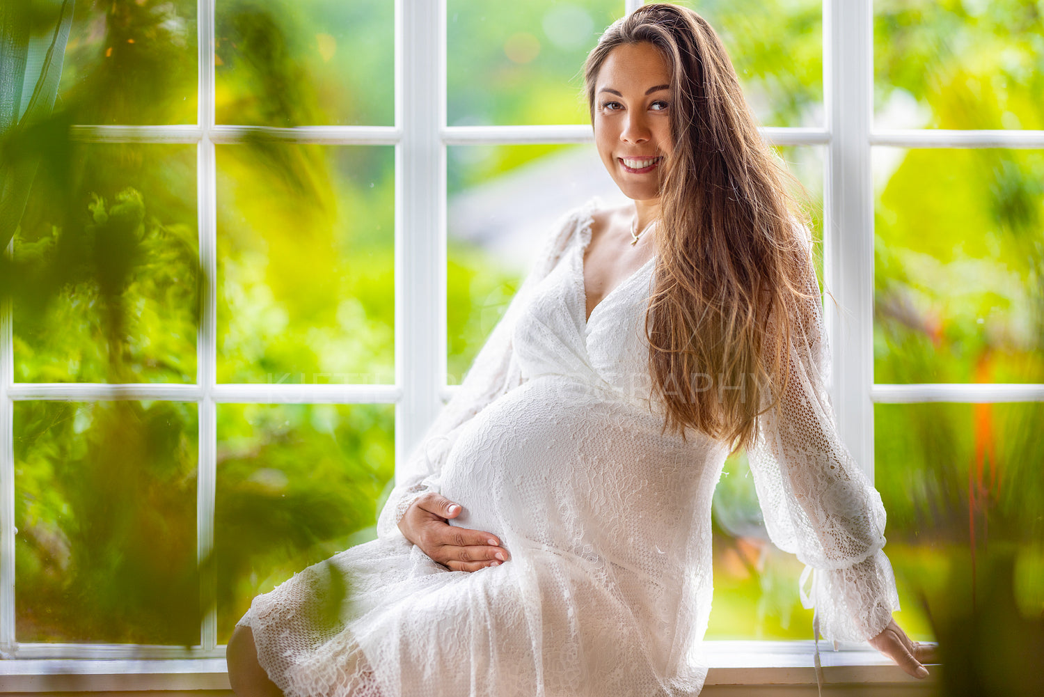 Smiling pregnant woman sitting in the window and holding her belly