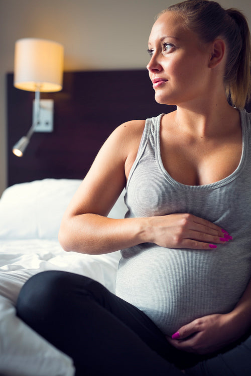 Pregnant woman sits on bed with her hand at belly
