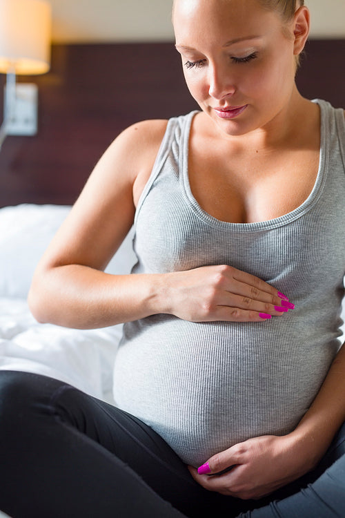 Pregnant woman sits on bed with her hands at belly