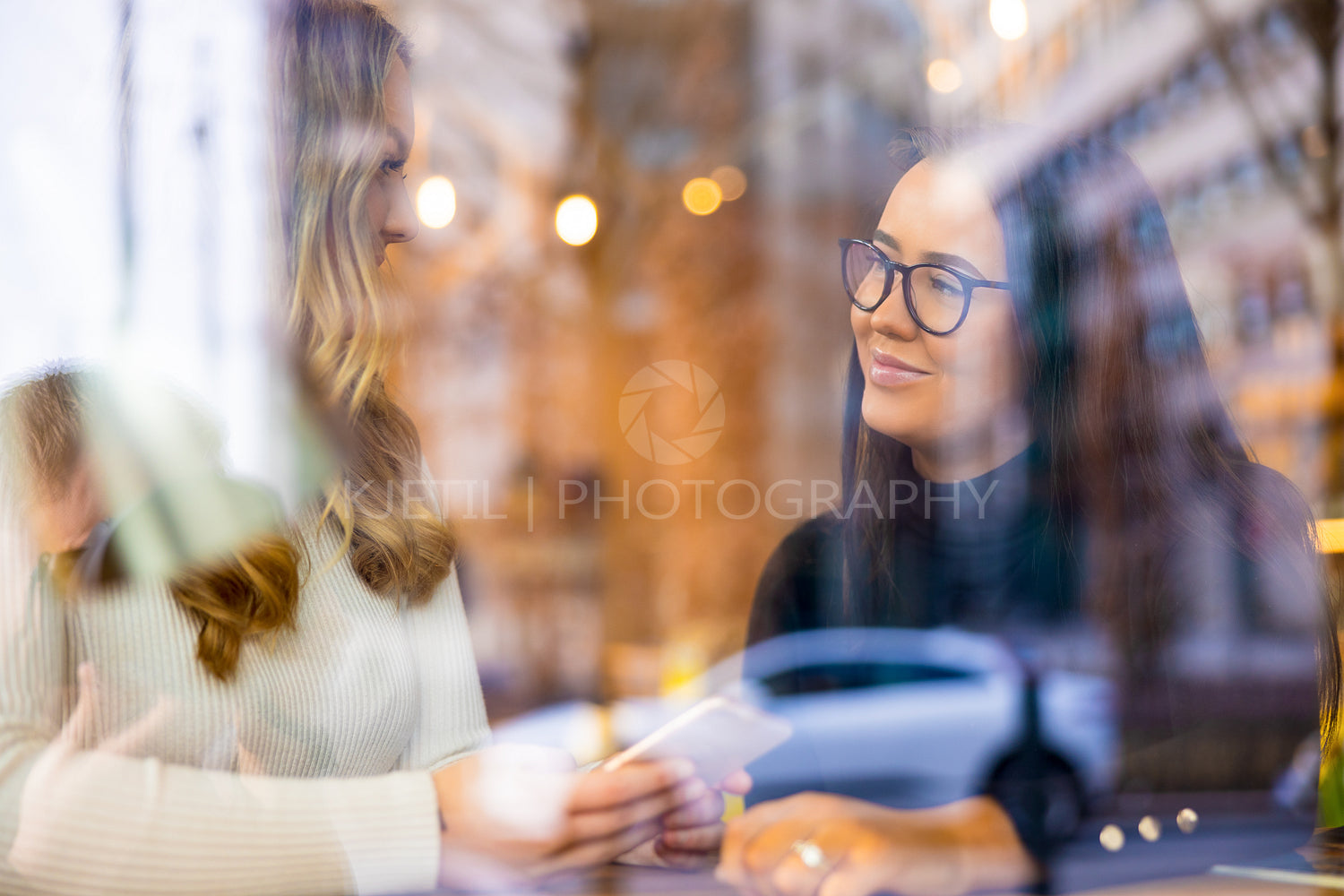Two Young Women Talking At Cafe in City Seen Through Window