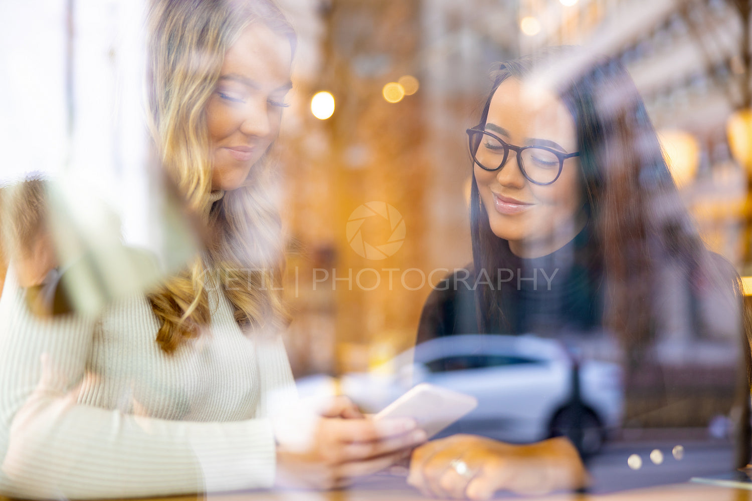 Two Smiling Friends Looking At Smartphone In Cafe