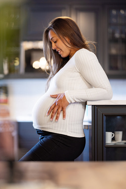 Expectant Smiling Mother Touching Her Pregnant Belly in the Kitchen