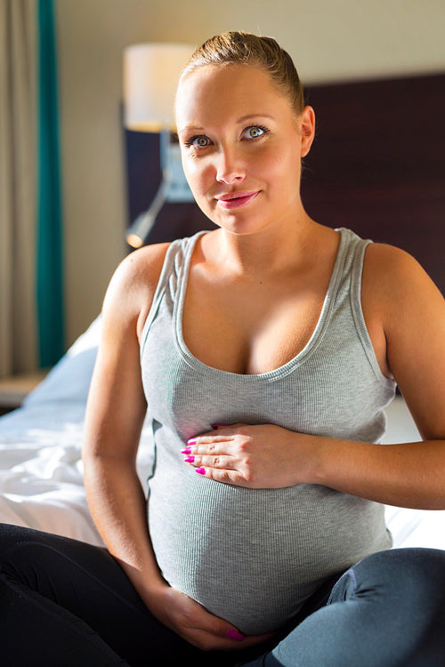 Smiling pregnant woman sits on bed with hand at belly