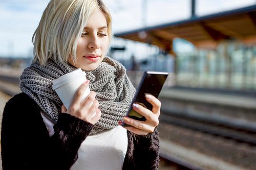 Woman With Coffee Cup Using Mobile Phone At Train Station