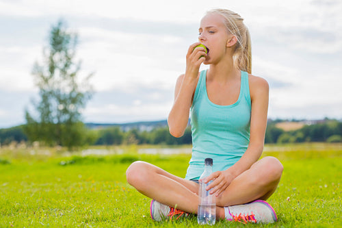 Woman sitting at the grass and eating apple after workout