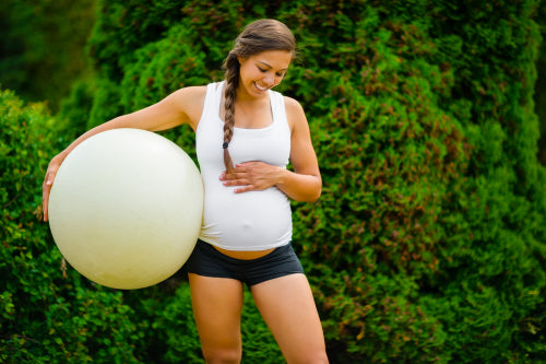 Expectant Mother Touching Stomach While Holding Exercise Ball In Park