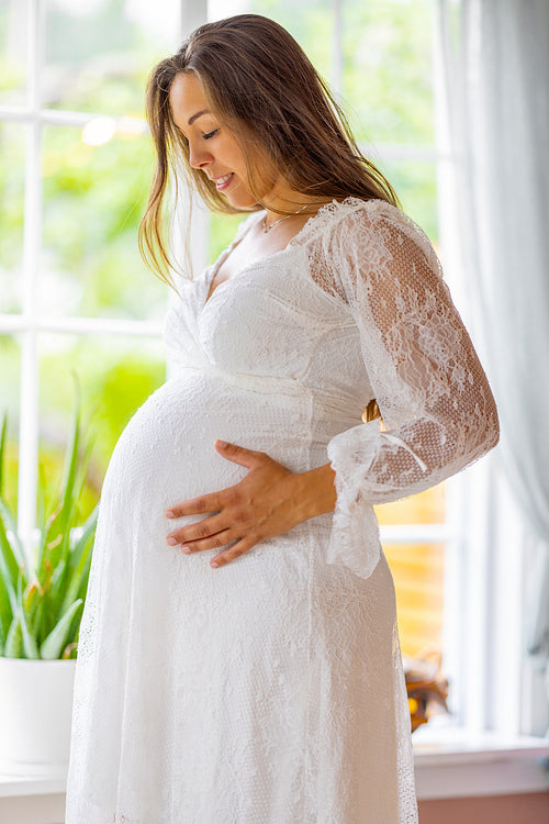 Smiling pregnant woman standing by the window and touching her belly