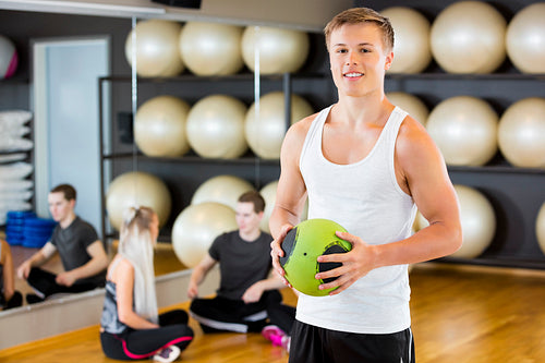 Confident Man Holding Medicine Ball While Friends Resting In Gym