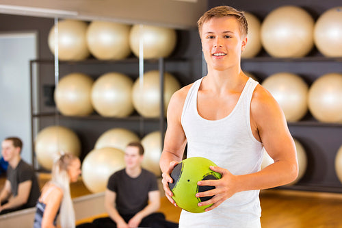 Smiling Man Holding Medicine Ball While Friends Resting In Gym