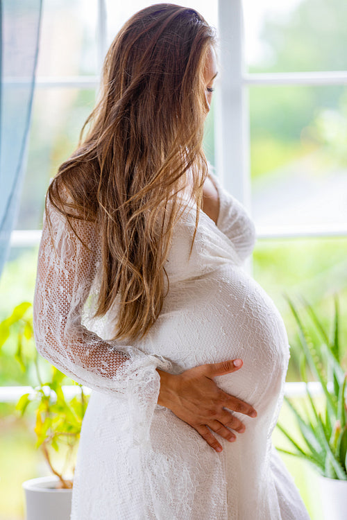 Pregnant woman standing by the window and touching her belly