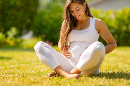 Pregnant Woman in White Sitting On Grass Outdoors Holding Belly